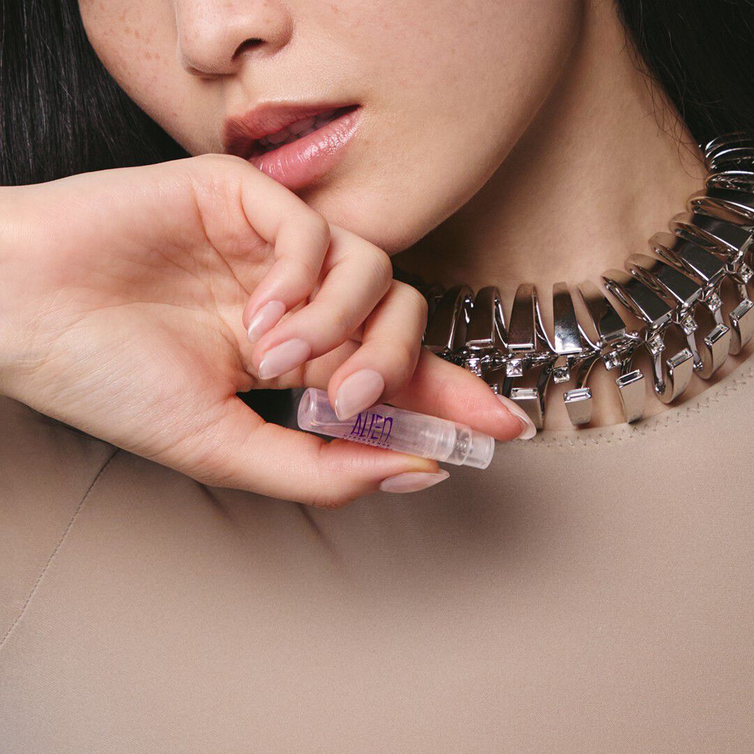 A close-up of a woman wearing a chunky metallic silver necklace and holding a small Mugler Alien perfume sample vial near her lips.