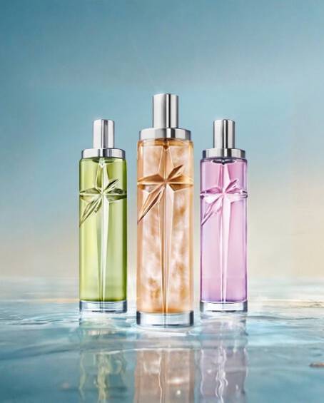 Three slender glass perfume bottles in green, gold, and purple standing on a reflective wet surface under a light blue sky.