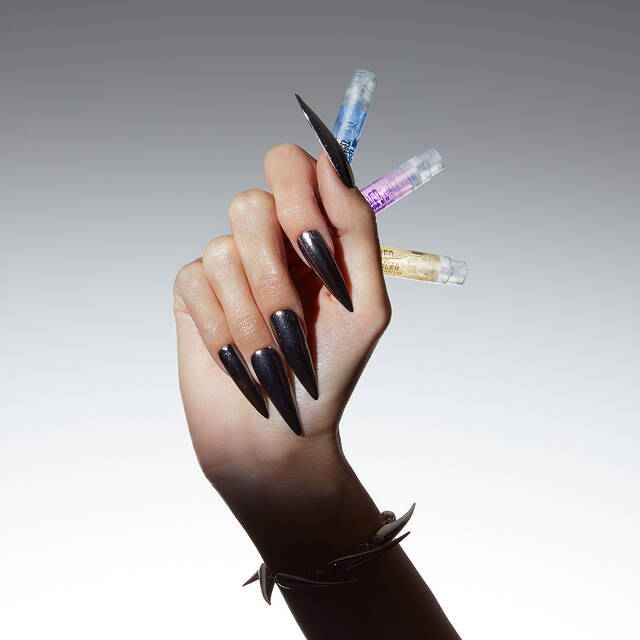 A hand with long, pointed black nails holds three small vials of colorful liquids against a light gradient background.