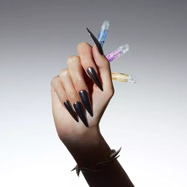 A hand with long, sharp black nails holding four small vials of different colors against a gradient background.