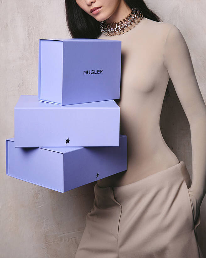 Model posing beside a stack of three purple Mugler boxes.