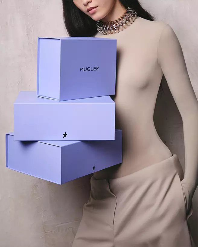 Model posing beside a stack of three purple Mugler boxes.
