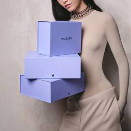 Model posing beside a stack of three purple Mugler boxes.