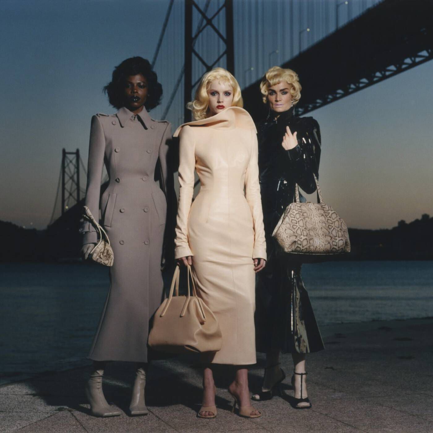 Three models in structured high-fashion outfits carry designer handbags on a waterfront walkway at dusk, with a suspension bridge in the background.