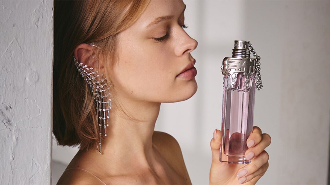 de-profile of a woman with closed eyes holding a tall, pink perfume bottle near her face, wearing a striking cascading rhinestone ear cuff.