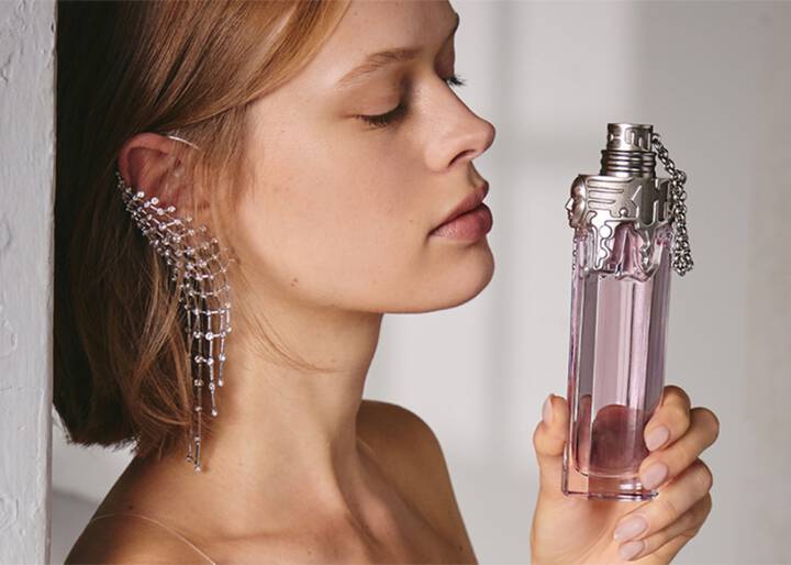 de-profile of a woman with closed eyes holding a tall, pink perfume bottle near her face, wearing a striking cascading rhinestone ear cuff.
