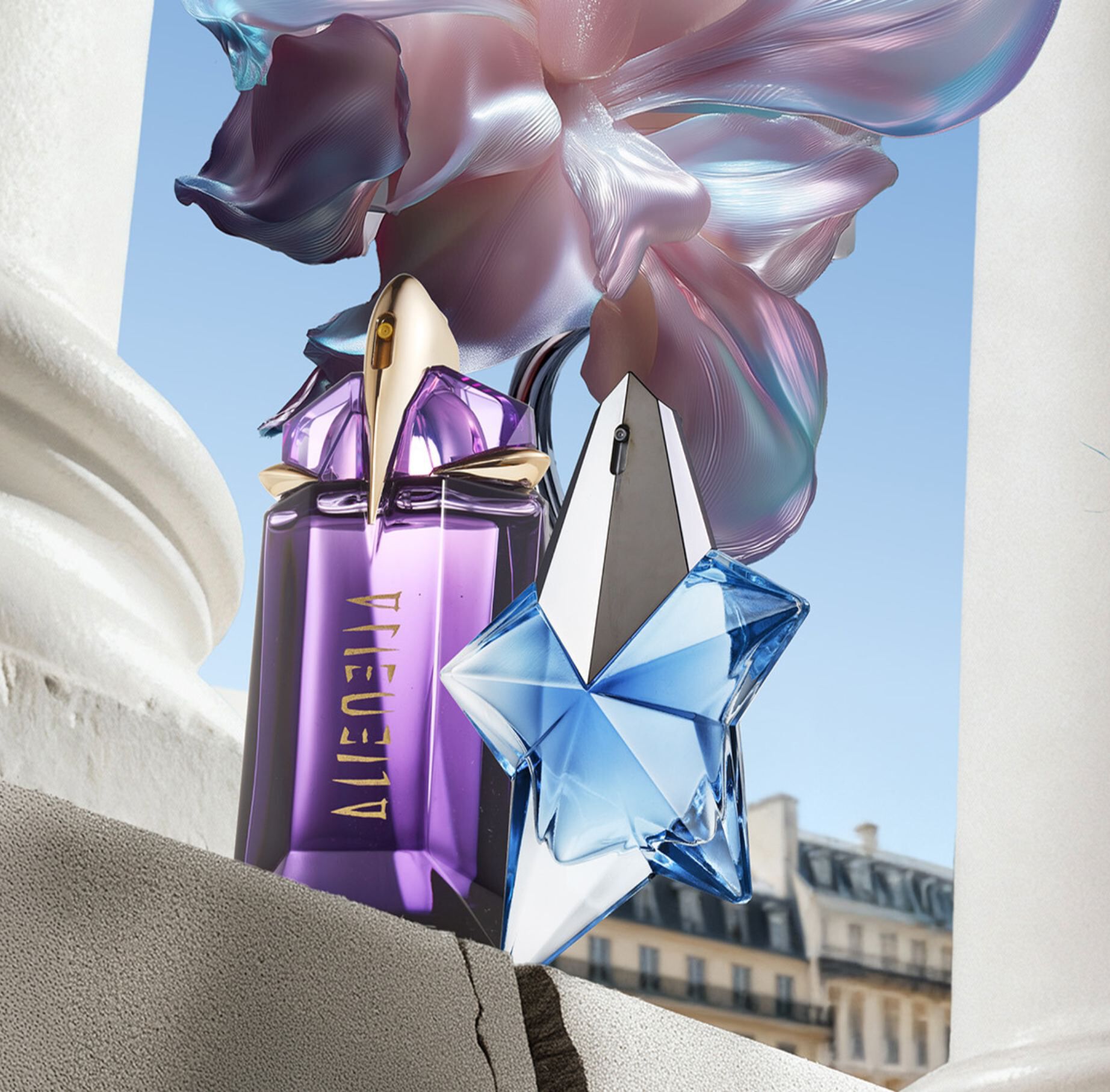 Mugler Alien and Angel perfume bottles displayed on a stone ledge against a Parisian cityscape and a large iridescent flower.