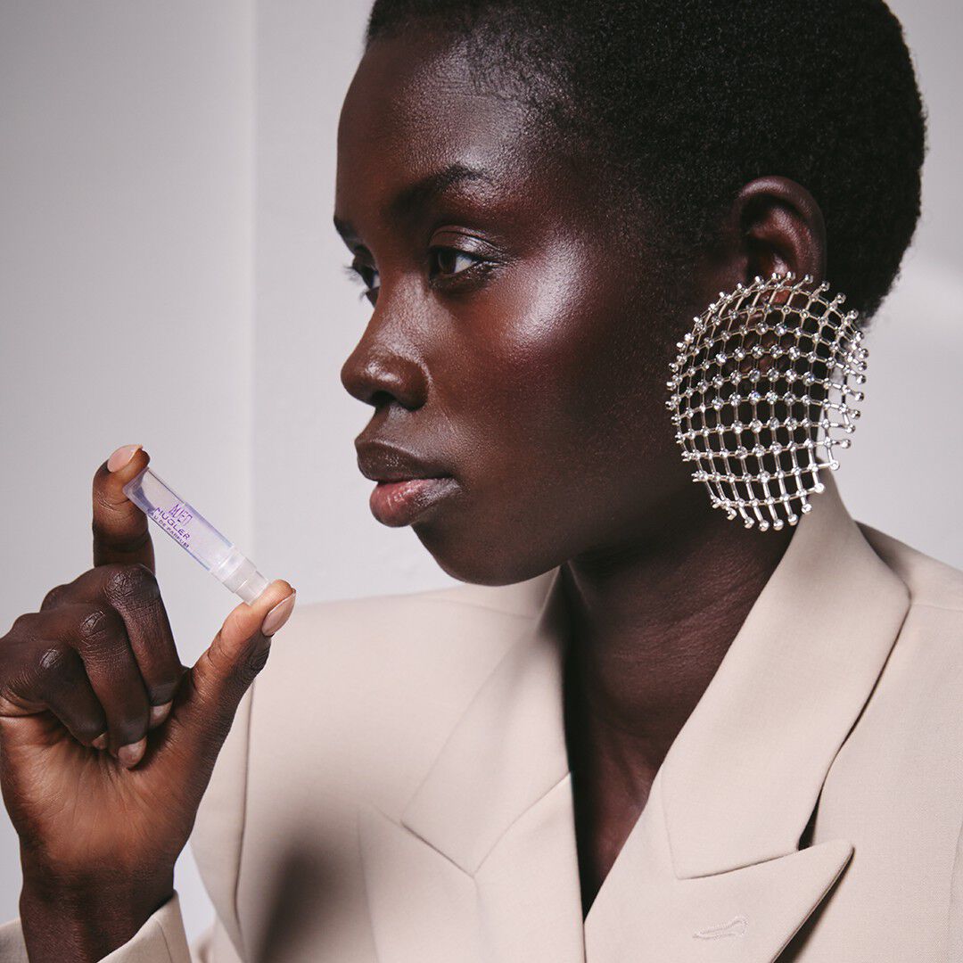 A profile of a Black woman with short hair wearing a beige blazer and a statement circular crystal earring, holding a small Mugler Alien perfume sample vial.
