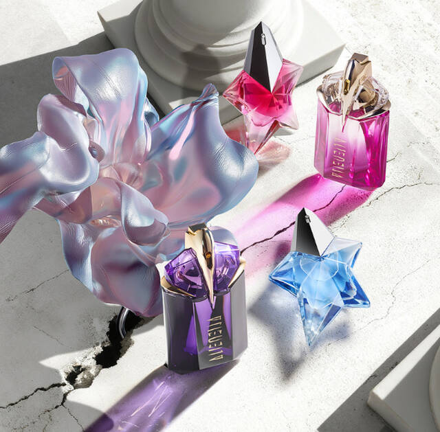 Four colorful Mugler perfume bottles in star and talisman shapes arranged on a cracked stone surface with an iridescent fabric flower