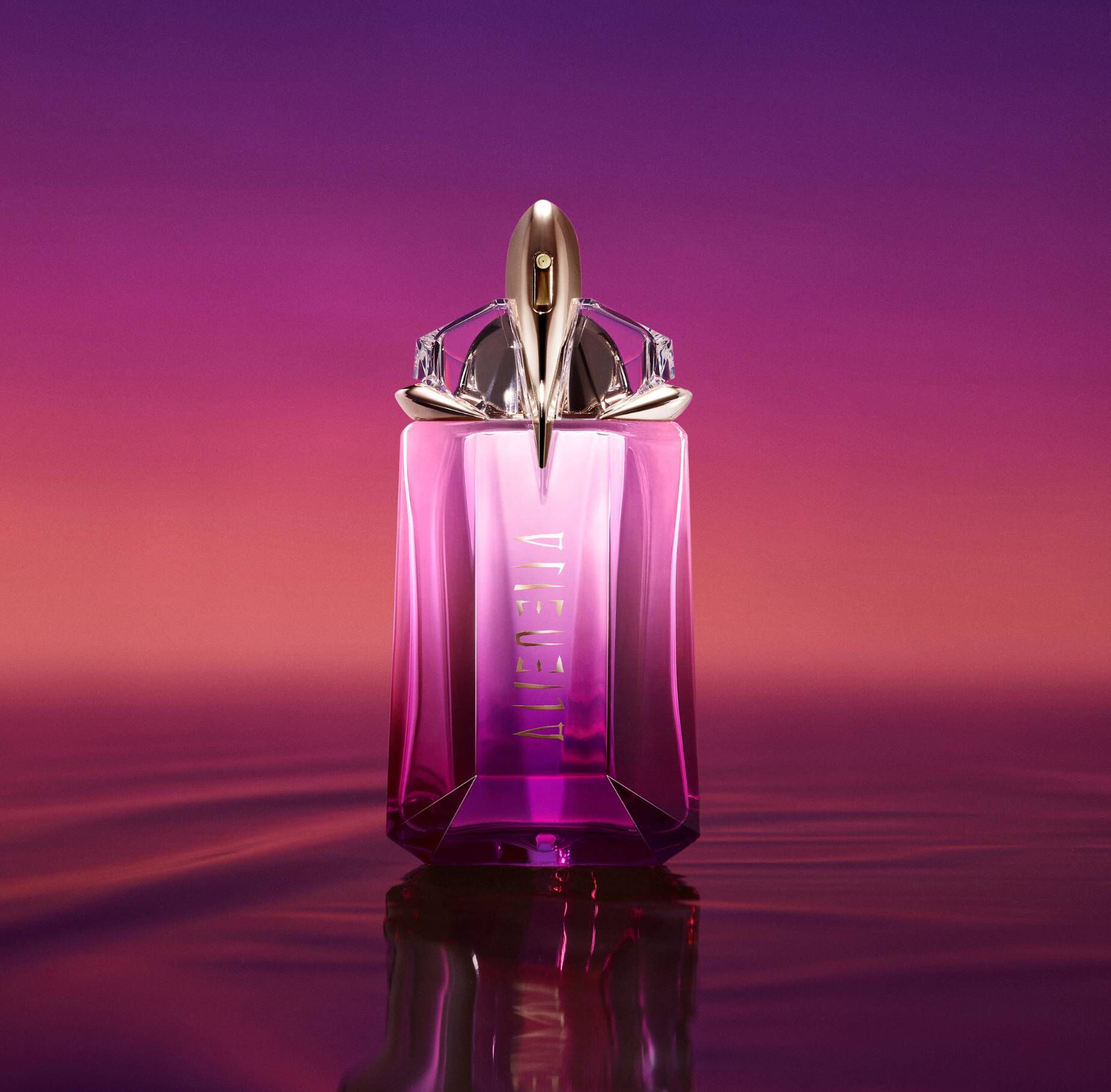 A bottle of Mugler Alien Hypersense perfume, featuring a vibrant pink and purple gradient glass bottle with gold accents, set against a matching sunset-colored background and reflecting in water.