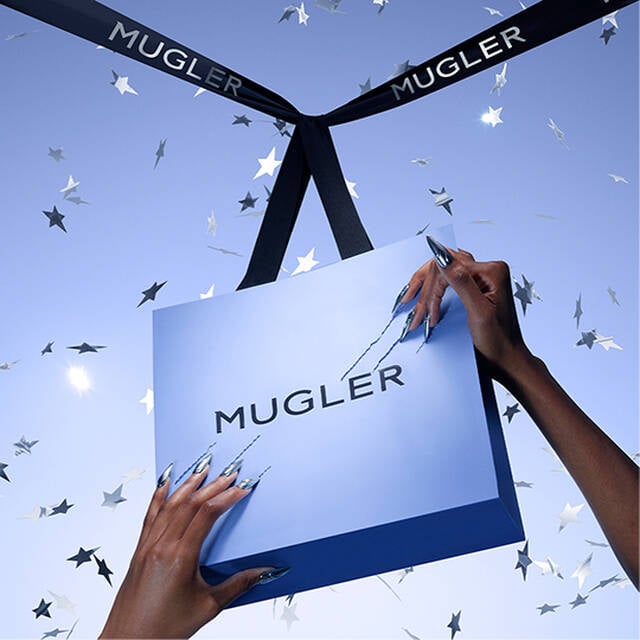 A light blue Mugler gift box is suspended with black ribbons, surrounded by silver star-shaped confetti. Hands with long, sparkling nails are holding the box, emphasizing its stylish presentation.