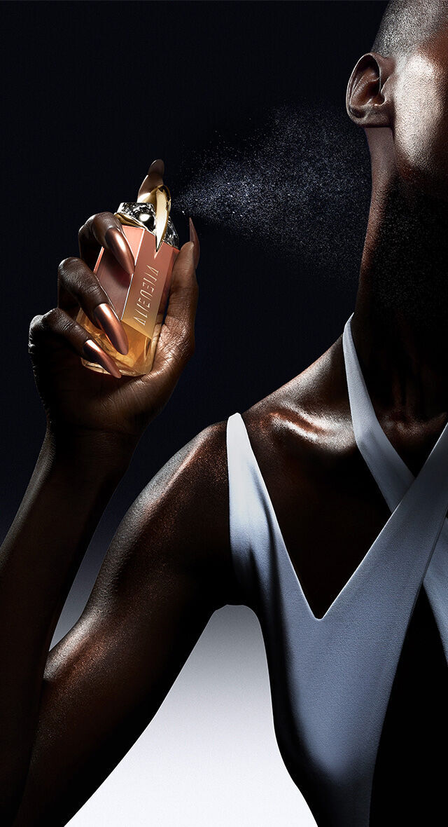 A close-up of a hand holding a stylish perfume bottle, spraying a fine mist against a dark background. The bottle features a sleek design with a luxury aesthetic.