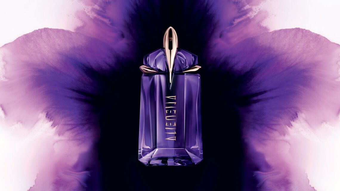 A futuristic purple and gold Mugler Alien perfume bottle displayed against an artistic watercolor-style purple flower bloom.