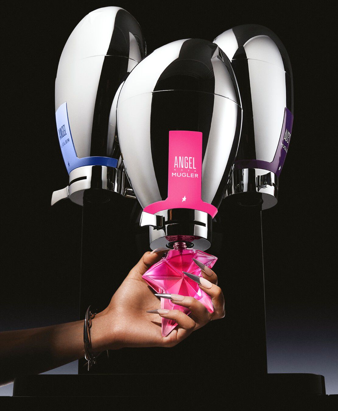 A photograph of three reflective, metallic perfume dispensers, one with a pink label, one with a blue label, and one with a purple label, on a dark background. A hand with long, pointed nails holds a pink star-shaped perfume bottle.