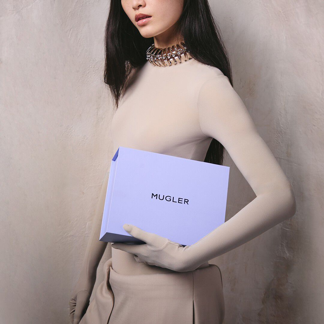 A close-up of a woman in a high-neck beige top and matching gloves holding a light purple 'MUGLER' brand box, styled with a chunky silver collar necklace.