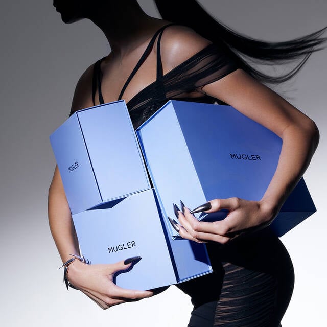 A silhouette of a person sitting with long hair, holding a white shopping bag with "MUGLER" printed on it, against a sleek reflective surface.
