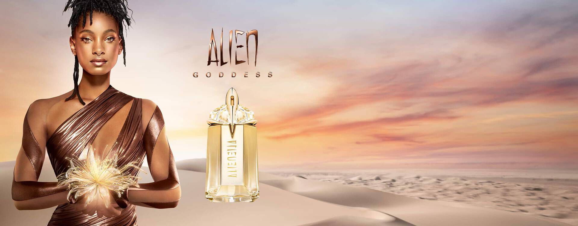 A model wearing a bronze dress poses with a bottle of Alien Goddess perfume against a desert landscape and sunset, showcasing elegance and allure.