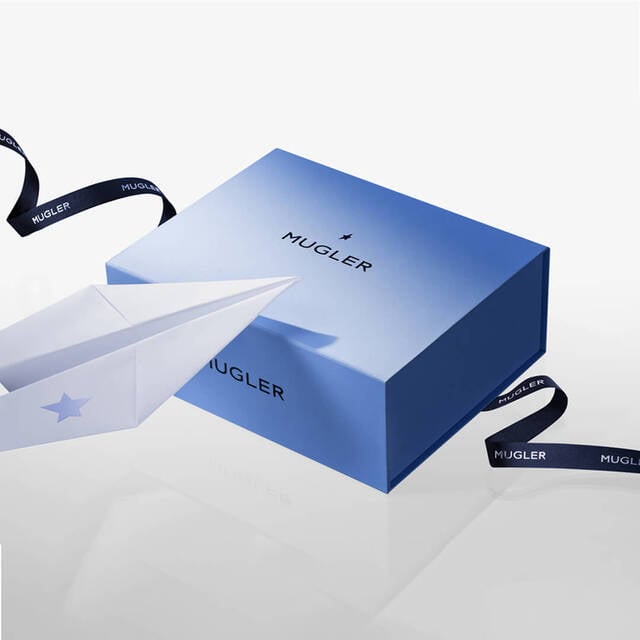 A blue gift box with the Mugler logo, featuring a white paper airplane nearby and black ribbon with the Mugler name.