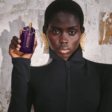 A Black woman with short hair wearing a black zip-up top and ornate gold statement earrings, holding a purple and gold Mugler Alien perfume bottle.
