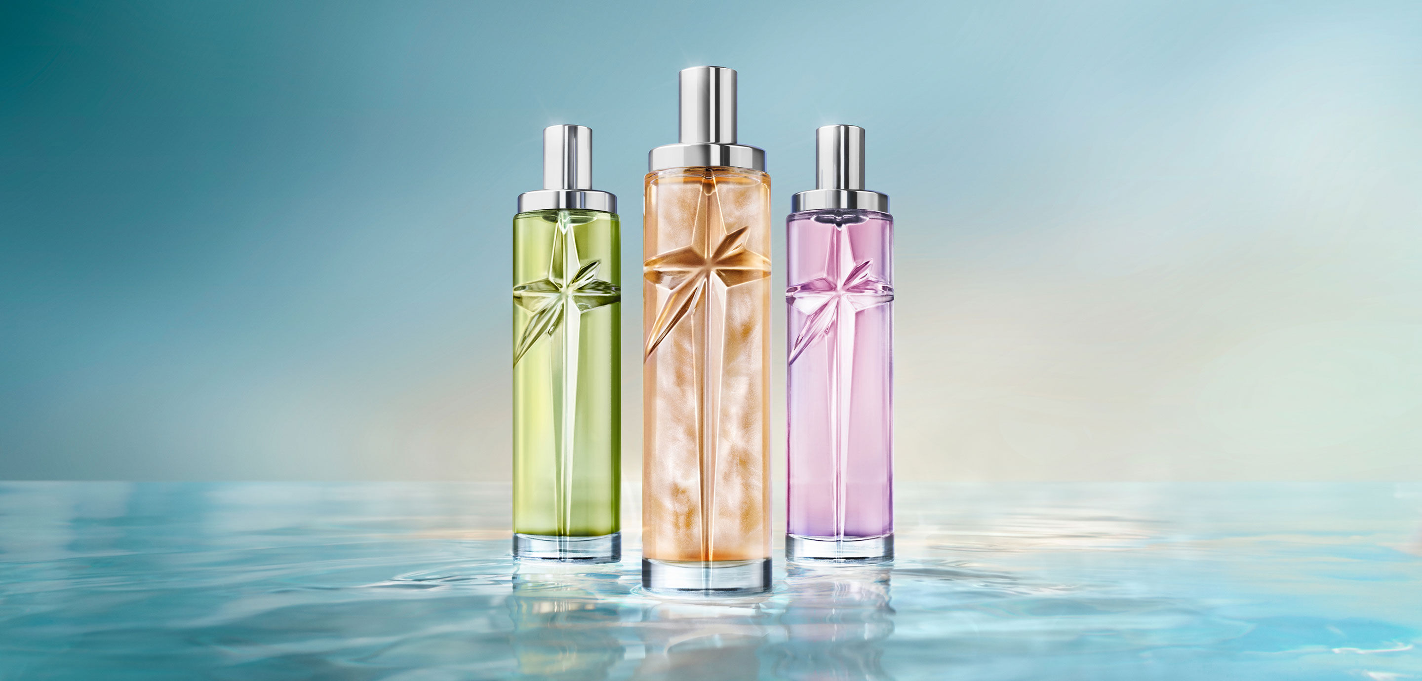 The new Starlicious collection: three tall cylindrical perfume bottles with silver caps in green, golden-amber and lavender-purple, standing side-by-side on a reflective water-like surface against a soft light blue background.