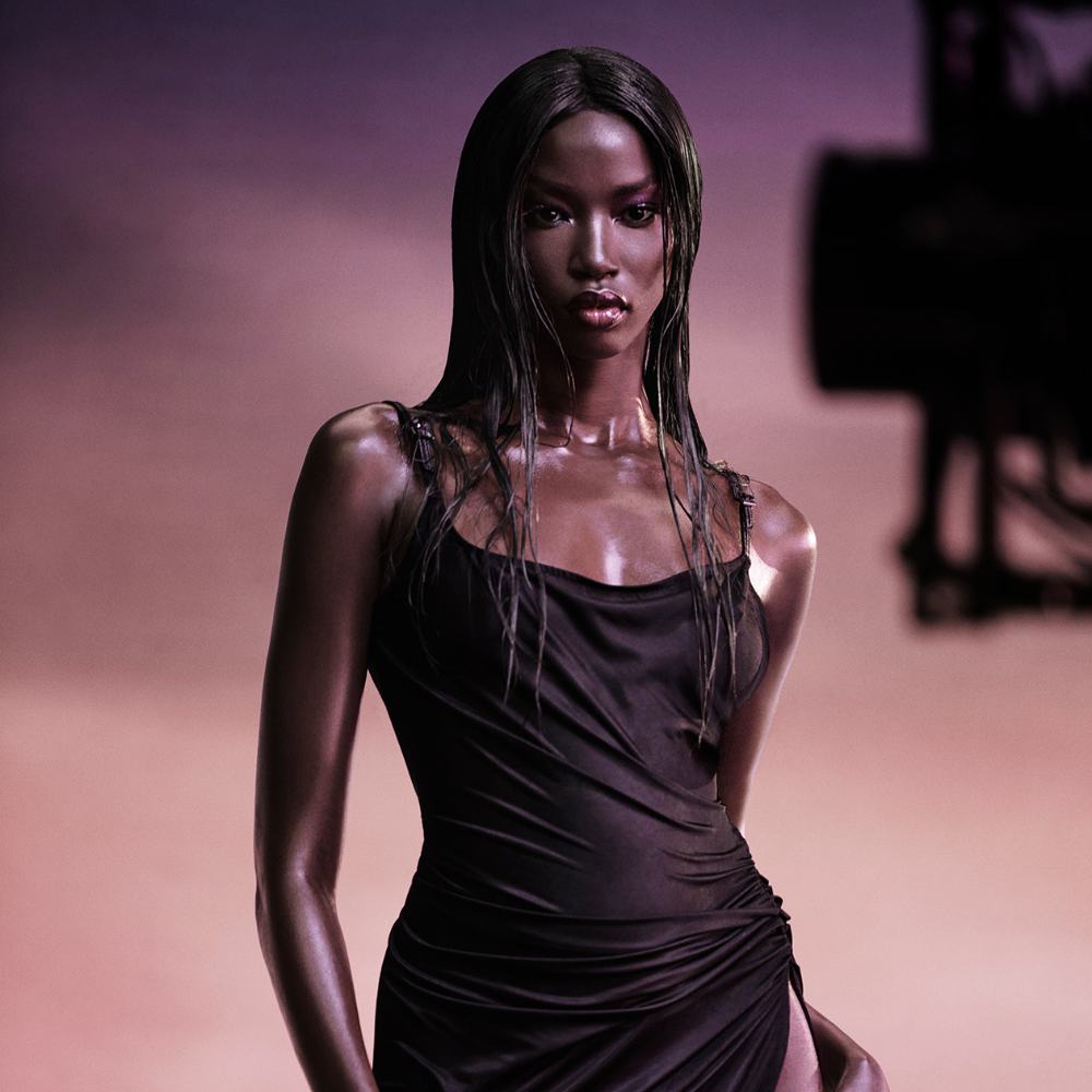 A model with dark, wet-look hair and shimmering skin, in a thoughtful pose, touching her face while looking away.