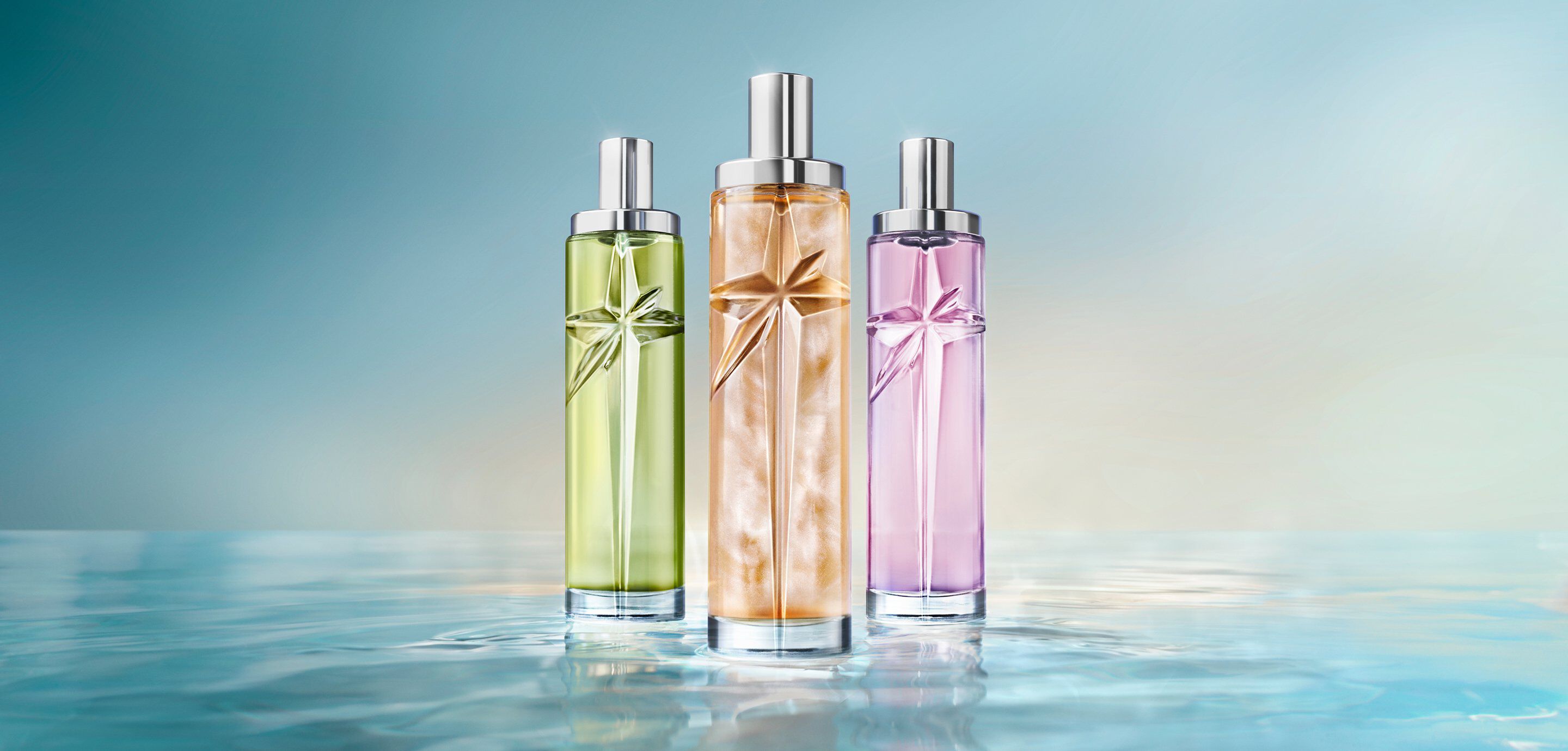 The new Starlicious collection: three tall cylindrical perfume bottles with silver caps in green, golden-amber and lavender-purple, standing side-by-side on a reflective water-like surface against a soft light blue background.