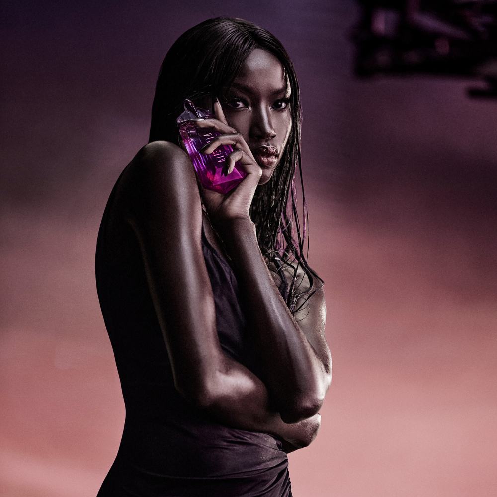 A striking model with dark, wet-look hair holds the vibrant pink Alien Pulp bottle delicately near her cheek, gazing intensely at the viewer, set against an ethereal purple and orange backdrop.