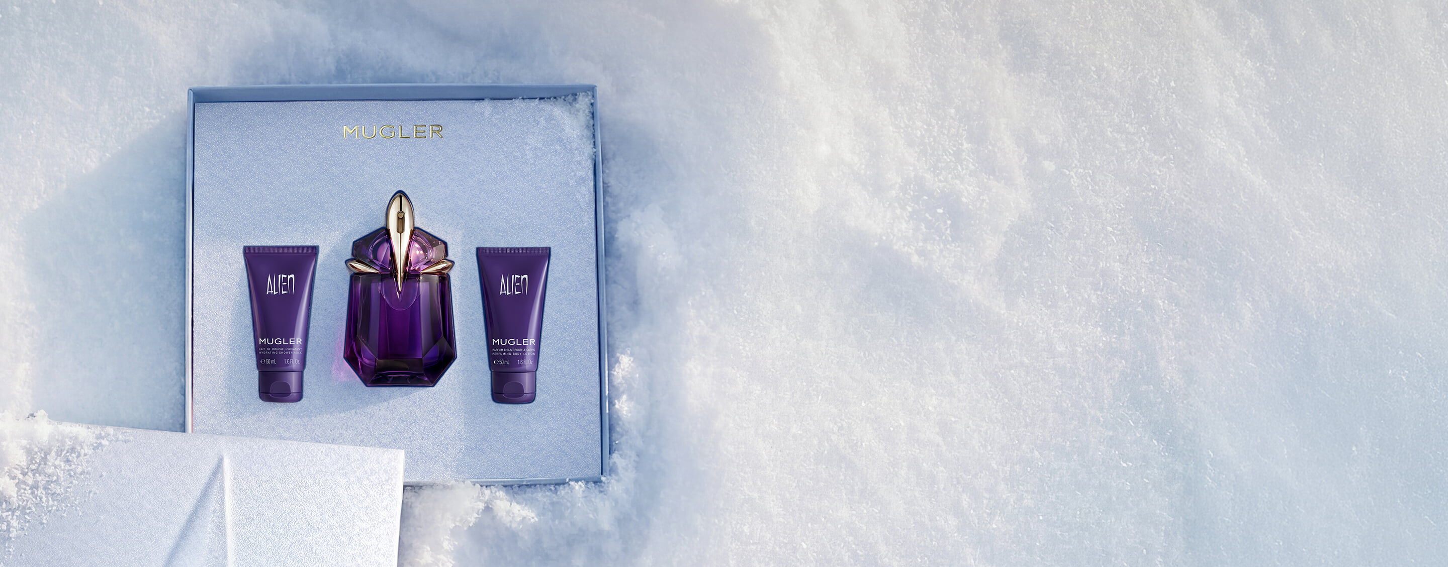 An open, textured ice-blue glistening Mugler gift set resting on snow, revealing an alien eau de parfum 30ml gift set. text reads "offer a gift to remember this holiday season, with alien eau de parfum's powerful floral notes wrapped in a shiny gift set"