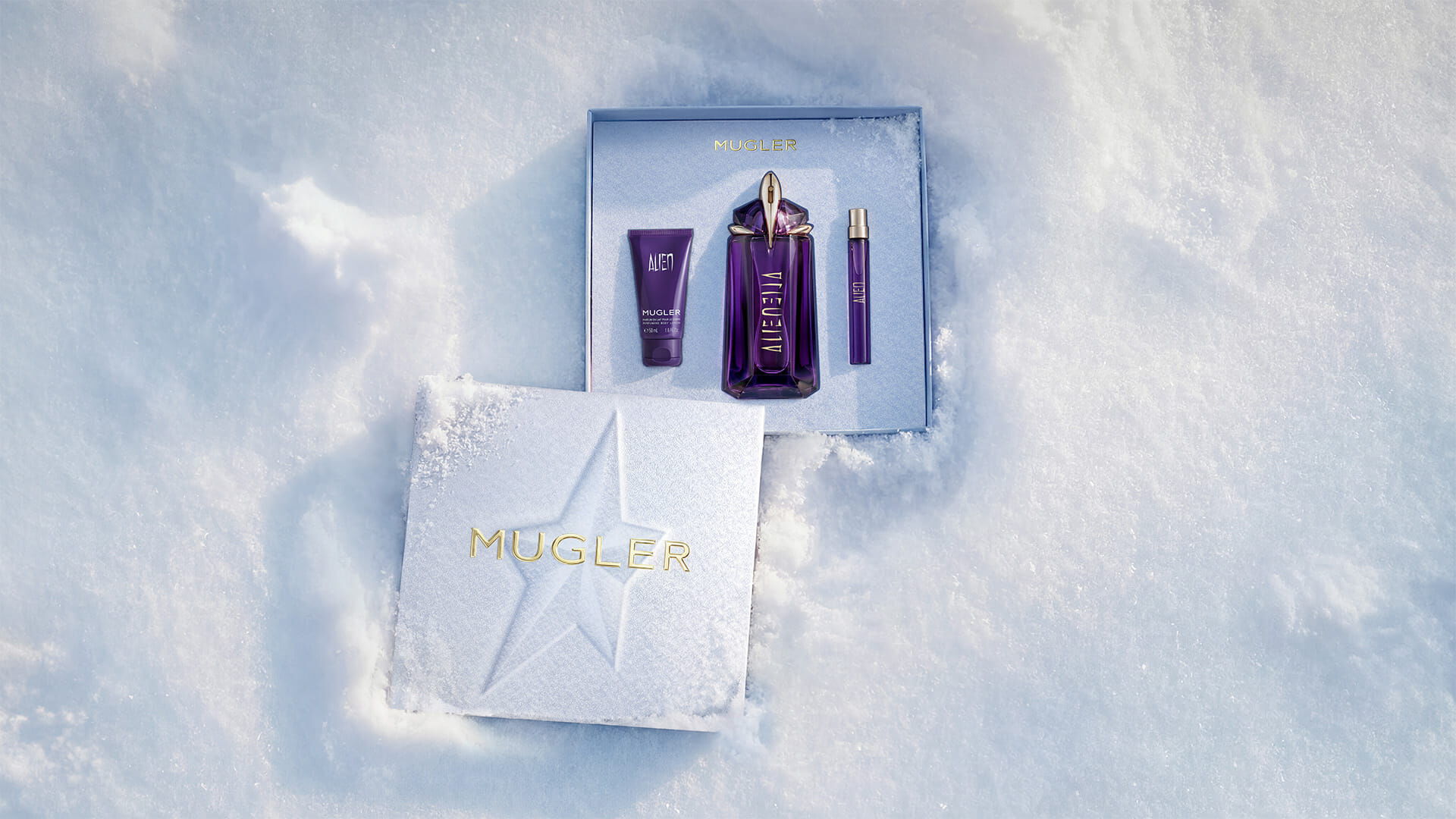 An open, textured ice-blue glistening Mugler gift set resting on snow, revealing an alien eau de parfum 60ml luxury gift set