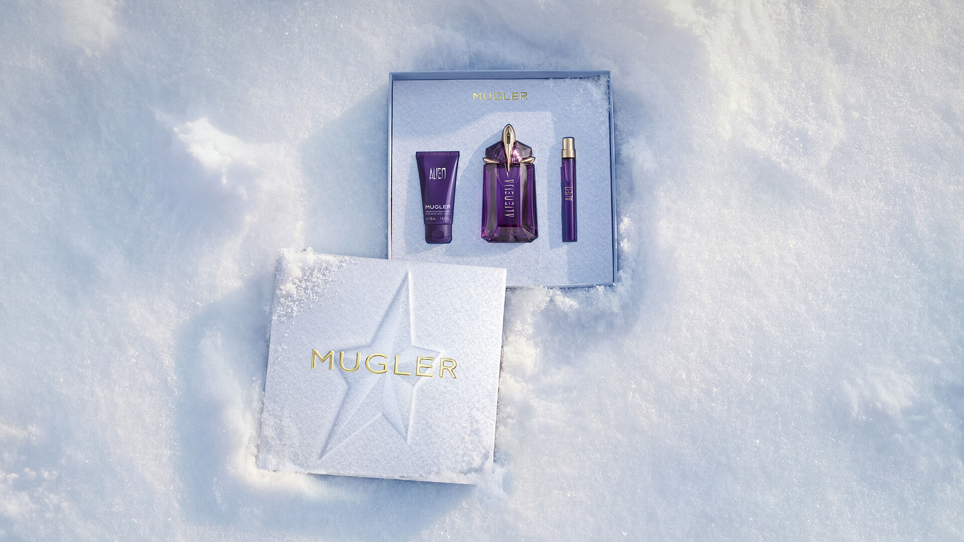 An open, textured ice-blue glistening Mugler gift set resting on snow, revealing an alien eau de parfum 60ml luxury gift set