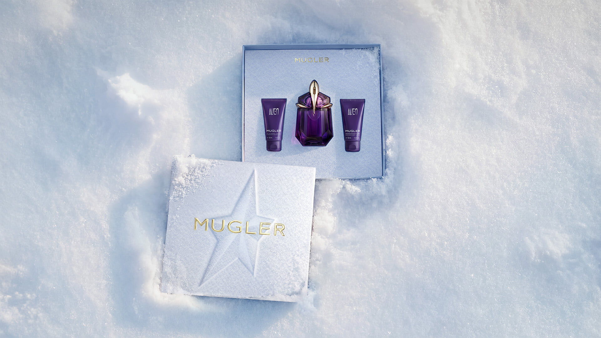 An open, textured ice-blue glistening Mugler gift set resting on snow, revealing an alien eau de parfum 60ml luxury gift set