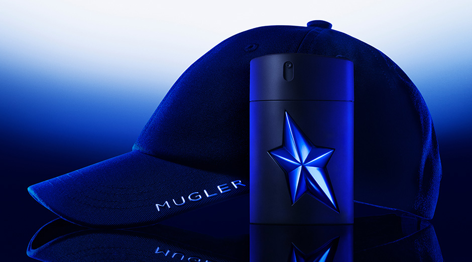 An A*Men Stellar Eau de Parfum bottle stands infront of a black Mugler cap, on a dark blue gradient background