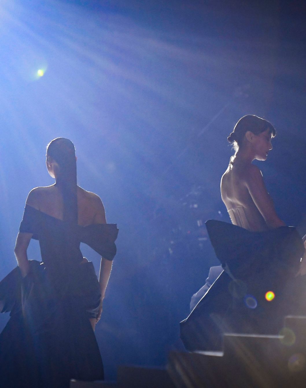 Two elegantly dressed models in dramatic black gowns pose back-to-back, illuminated by ethereal lighting on a stage.
