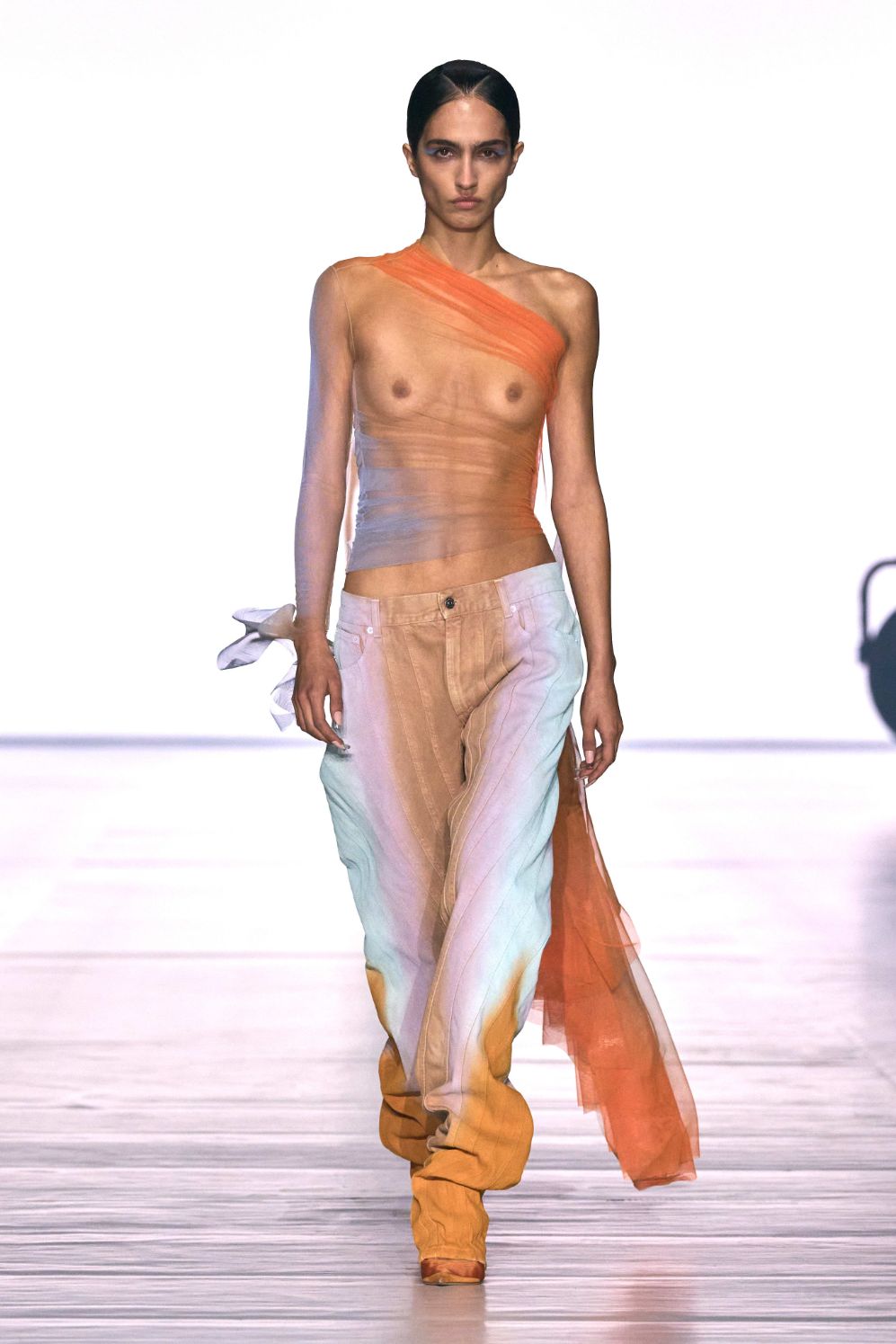 A model walks the runway in a sheer, colourful top and gradient trousers at a fashion show.