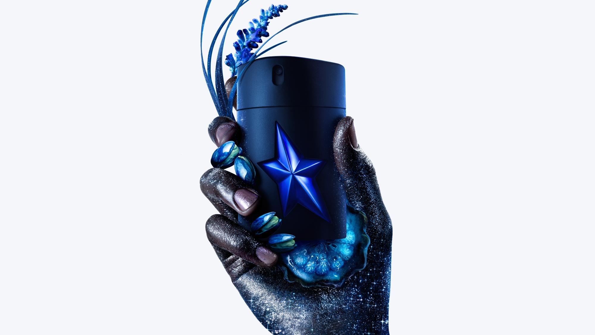 A hand is holding an A*Men Stellar Eau de Parfum bottle, with the key ingredients spilling from the bottle leaving a glittery trail.