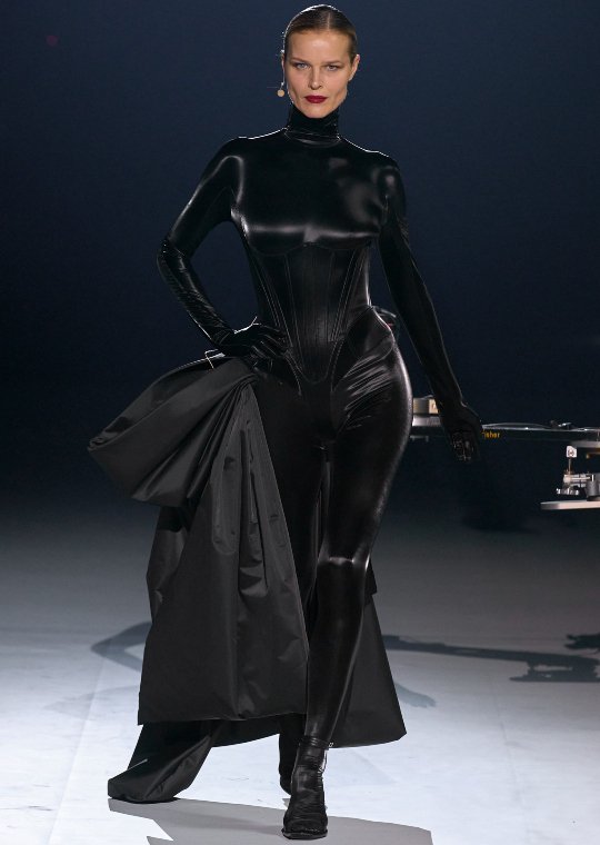 A model walks confidently on the runway in a sleek black bodysuit, holding a large black coat with a dramatic flair.