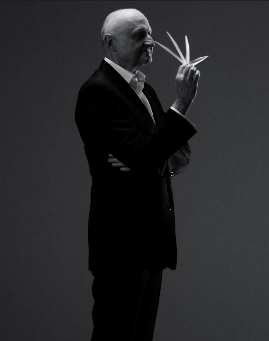 A poised older man in a black suit holds a pair of scissors, standing confidently against a dark backdrop.
